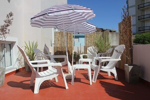 Un patio con sillas de plástico blancas y un paraguas a rayas, rodeado de plantas y una cerca de bambú.