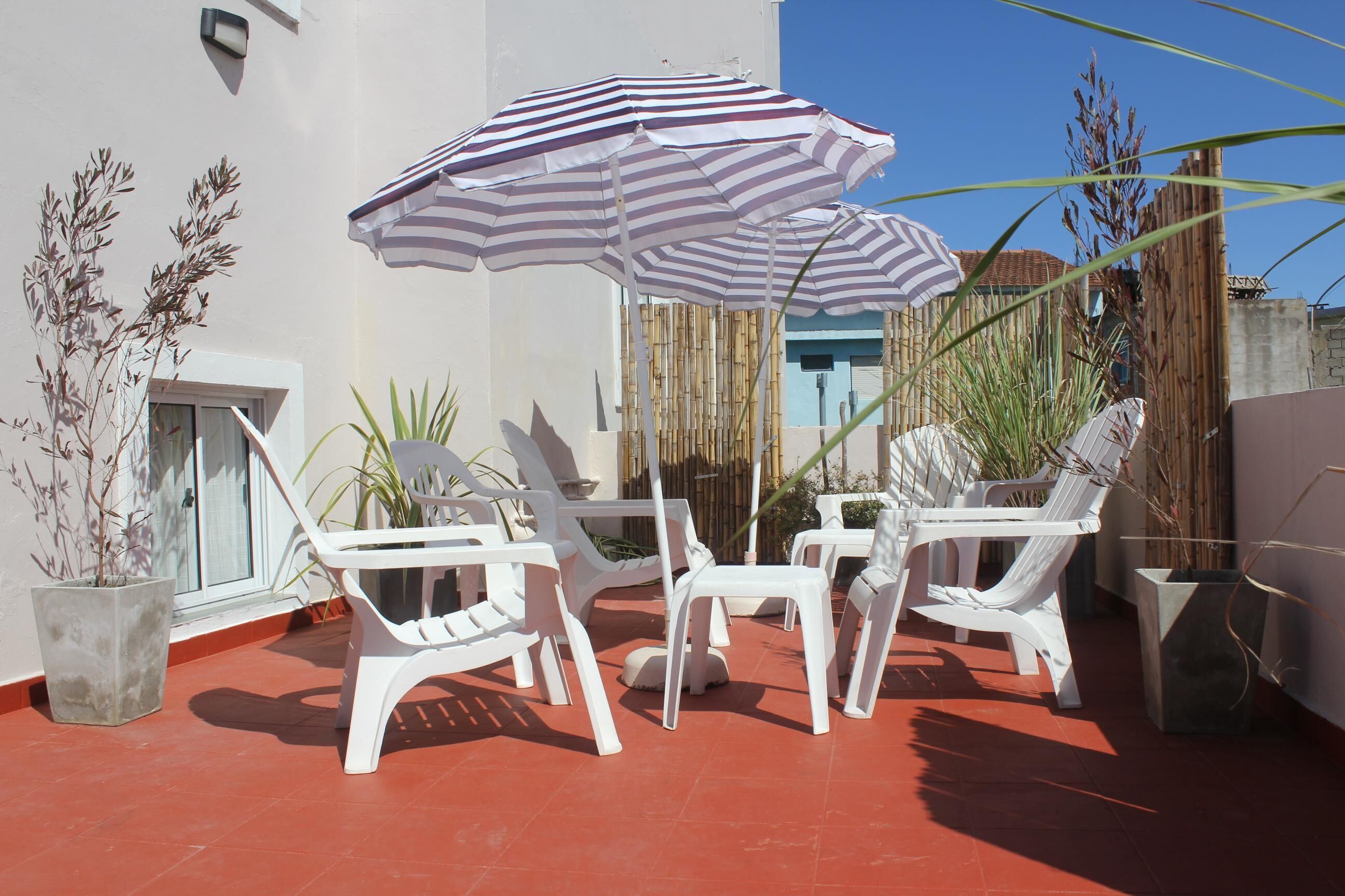 A patio with white plastic chairs and a striped umbrella, surrounded by potted plants and a bamboo fence.