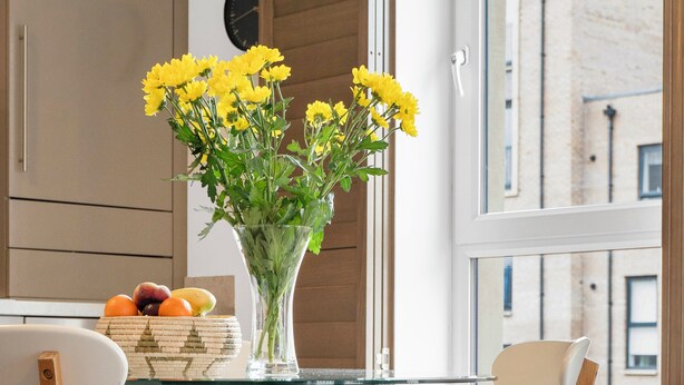 A modern kitchen with a glass table, wooden chairs, a vase of yellow flowers, and a fruit basket.