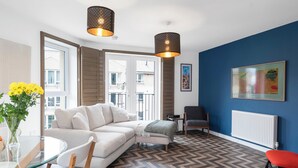 A modern living room with a white sofa, a glass table, and a red chair. There are two pendant lights, a radiator, and a blue wall with a framed picture.