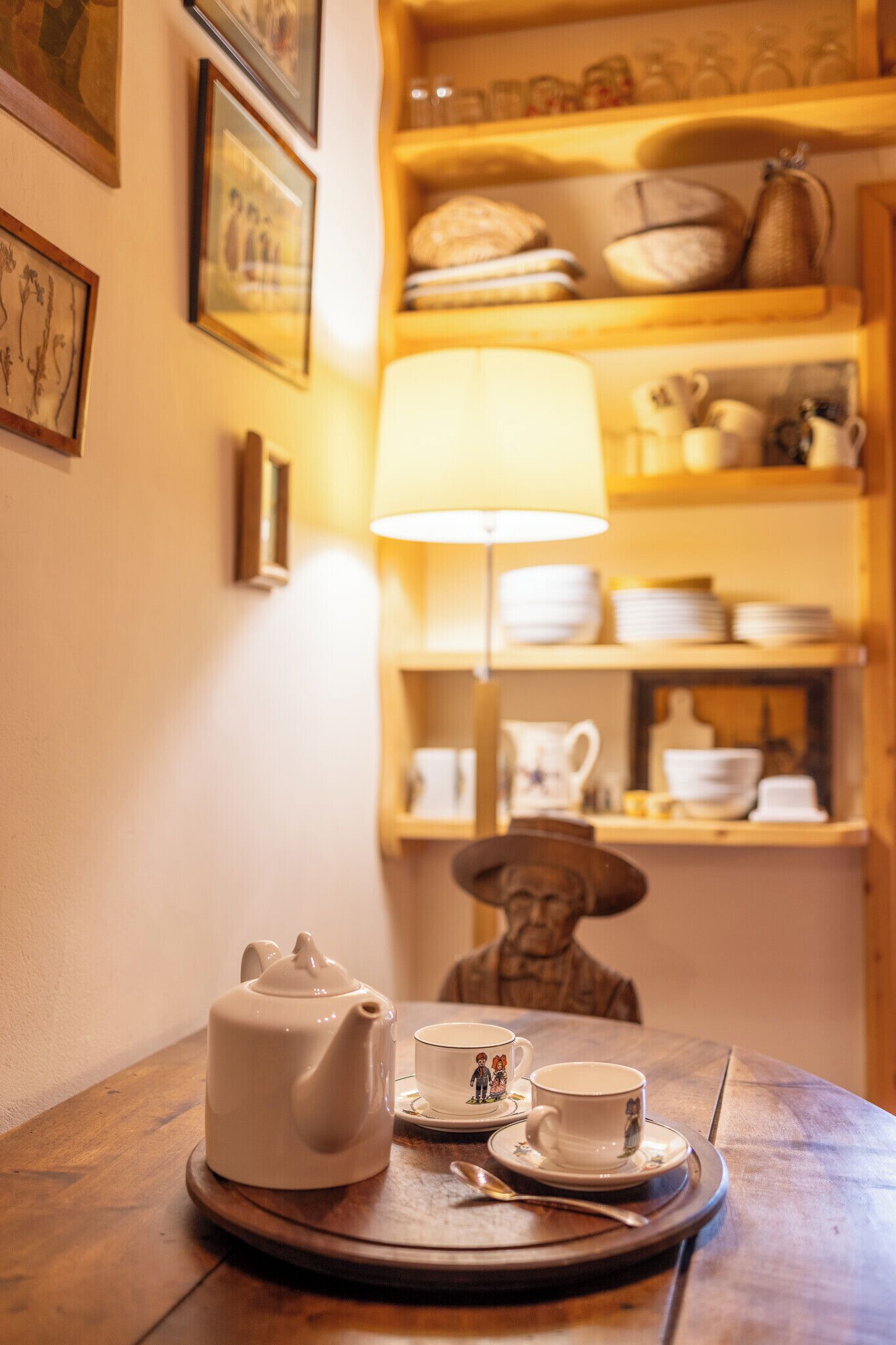 Une table avec une théière et des tasses, une lampe, et des étagères remplies de vaisselle et de poteries en arrière-plan.