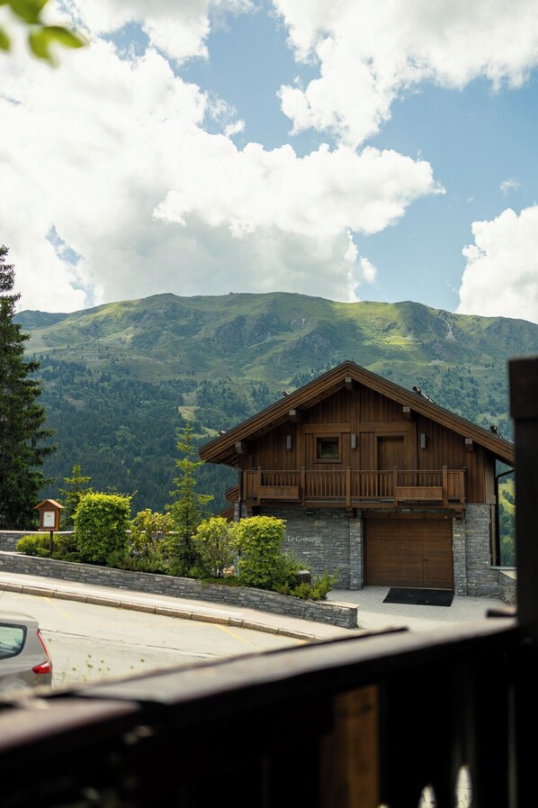 A wooden chalet with a stone foundation, set against a backdrop of lush green mountains and a partly cloudy sky.