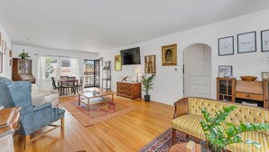 A spacious living room with a blue sofa, a wooden coffee table, and a patterned rug. There is a dining area with a table and chairs, and a wooden cabinet with a television on the wall. - Enchanting 2BR in Old Southwest (Reno)