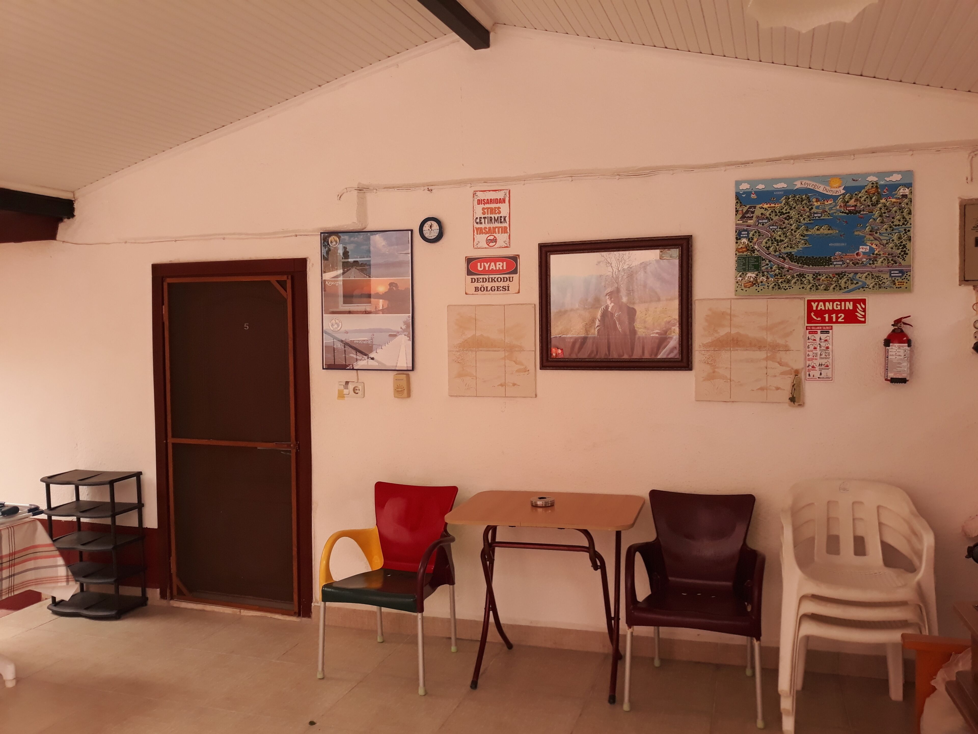 Una habitación con puerta, una mesa, sillas, una estantería y varios adornos en la pared, incluyendo un mapa y un extintor.