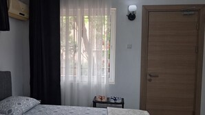 Ein Schlafzimmer mit einem Bett, einem Fenster mit durchsichtigen Vorhängen, einem kleinen Tisch mit Schuhen und einer Holztür.
