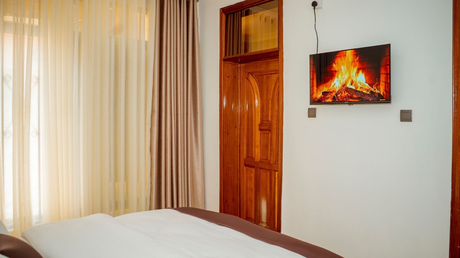 Une chambre à coucher avec un lit, une porte en bois et un téléviseur fixé au mur qui diffuse une image de cheminée.