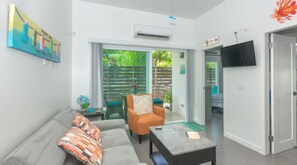 A modern living room with a sofa, a coffee table, and a television. There is a sliding glass door leading to an outdoor area with plants.