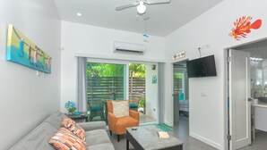 Een moderne woonkamer met een grijze bank, een oranje fauteuil en een salontafel. Er is een televisie aan de muur bevestigd en een plafondventilator.