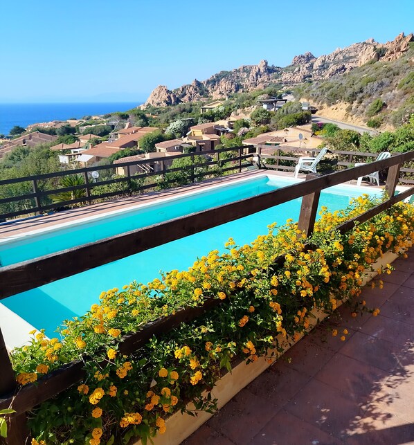 Une piscine avec une terrasse en bois et des fleurs jaunes au premier plan, entourée d’un paysage montagneux et d’un ciel dégagé.