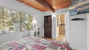 Ein Schlafzimmer mit einem großen Bett, einem Fenster mit Jalousien, einer Holzbalkendecke, einem Fernseher und einer Tür zu einem weiteren Raum.