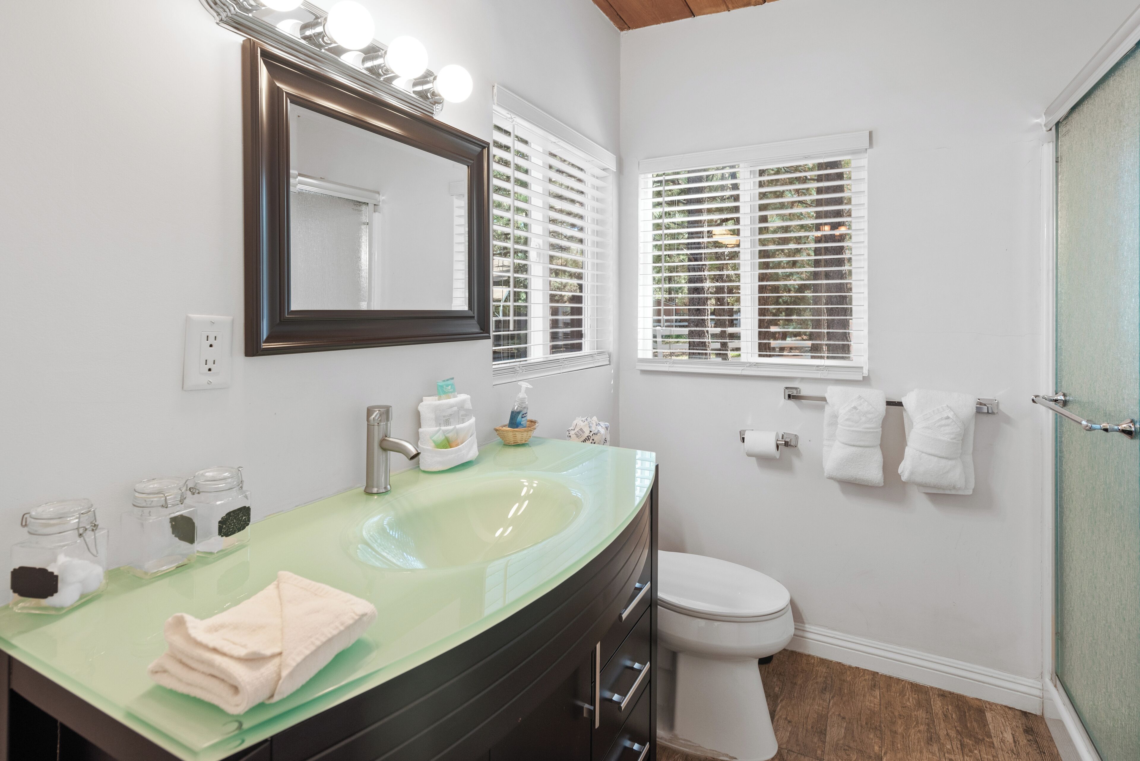 Un baño con un lavamanos de vidrio verde, un espejo grande, inodoro blanco y una ventana con persianas.
