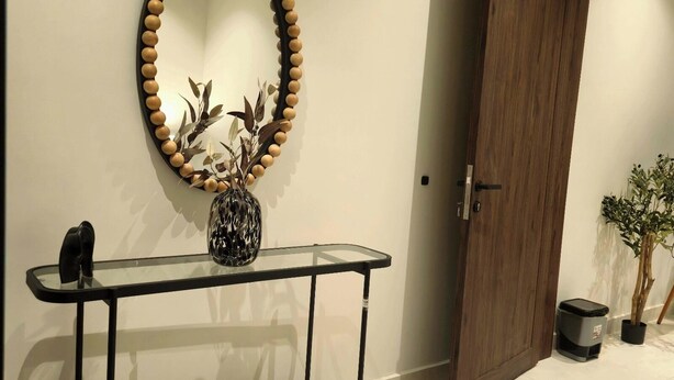 A hallway with a glass console table, a wooden bench, a mirror with a wooden frame, and a door. -  فايان استديو مودرن بسرير مزدوج وصالة ودخول ذكي في المنطقة الحية في شمال بريدة (Buraydah)