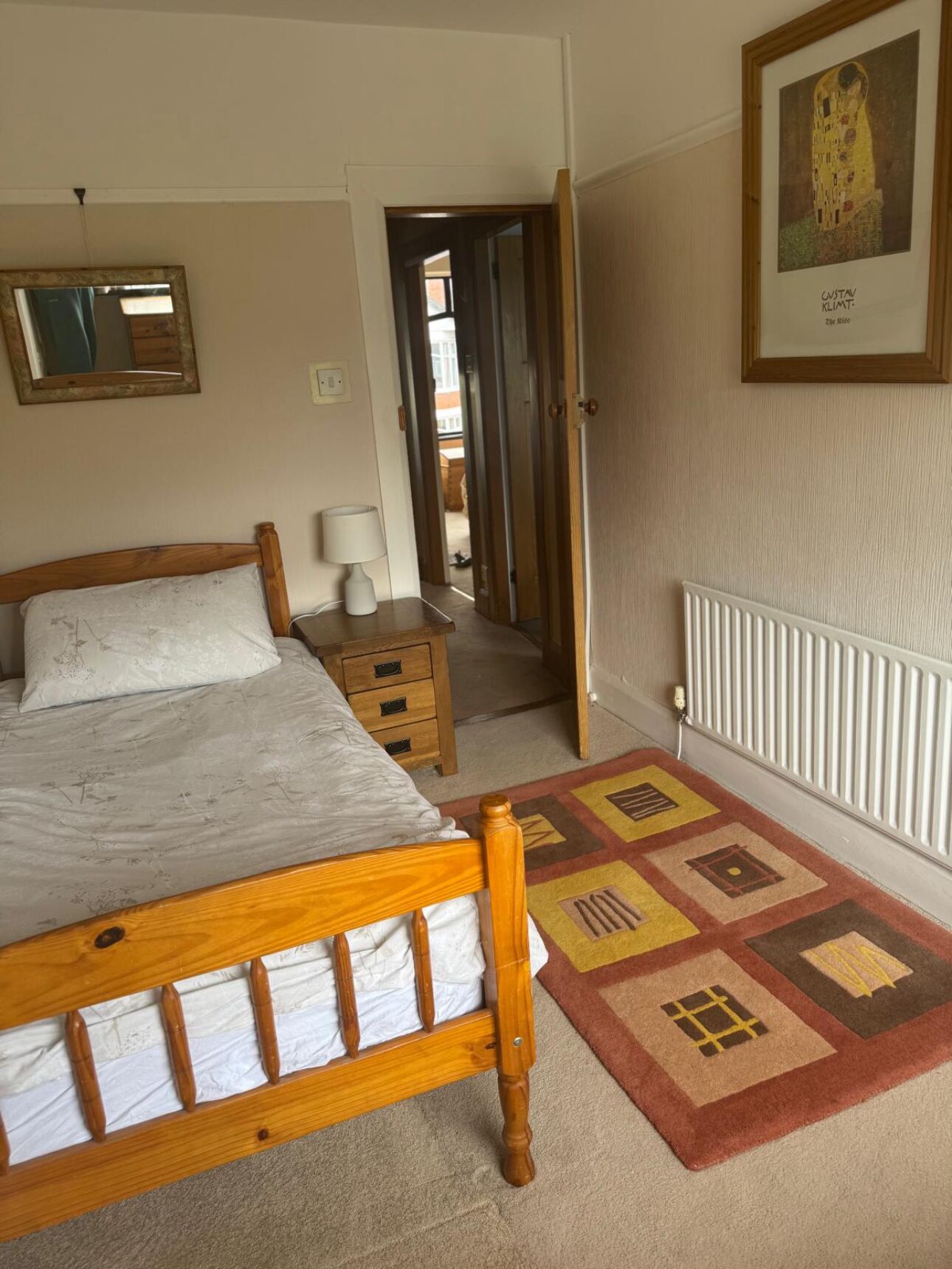 Quarto com cama de madeira, mesa de cabeceira, luminária, radiador, tapete estampado, quadro emoldurado e espelho.