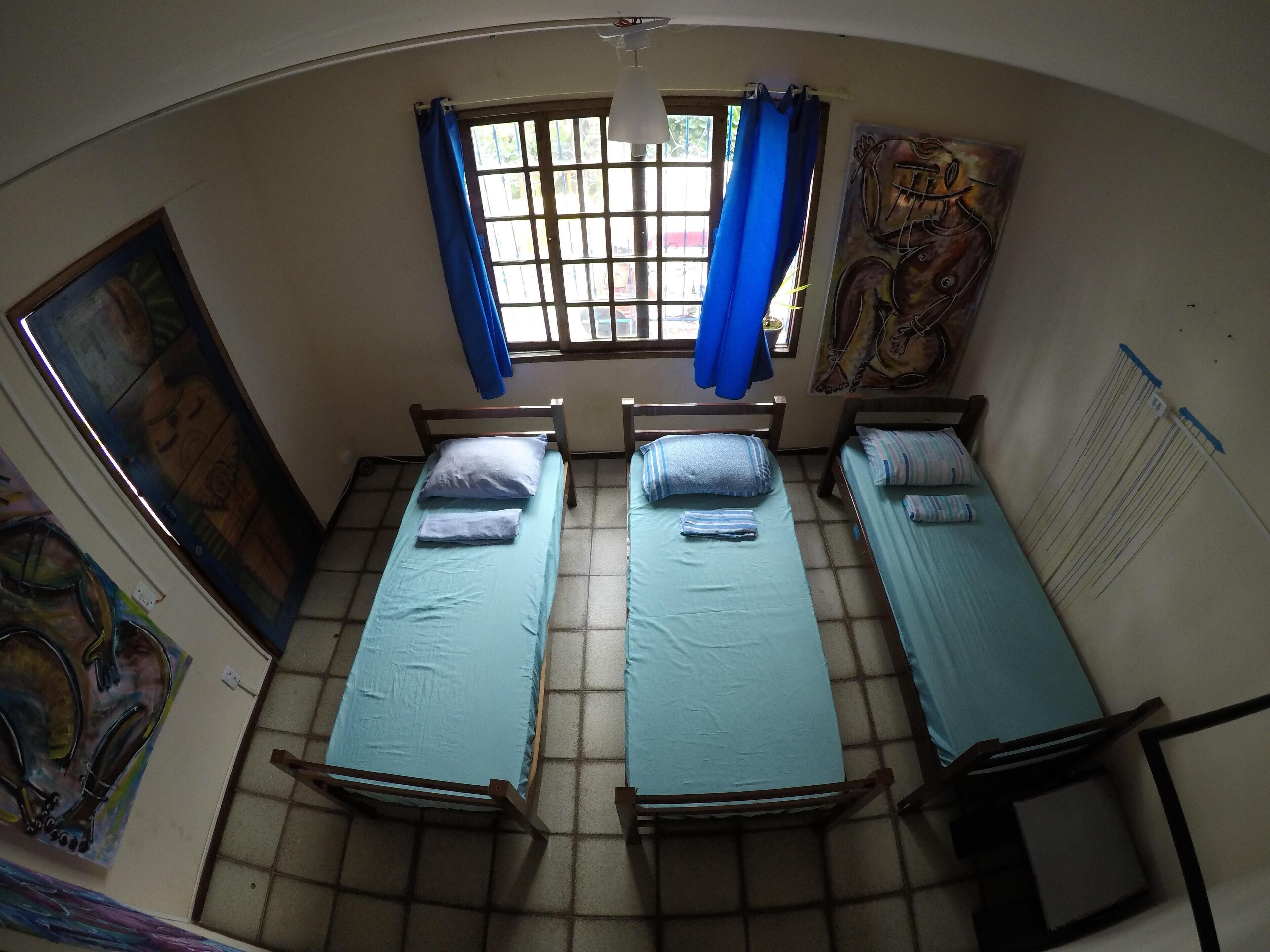 Een studentenkamer met twee eenpersoonsbedden, een raam met blauwe gordijnen en kunstwerken aan de muur.