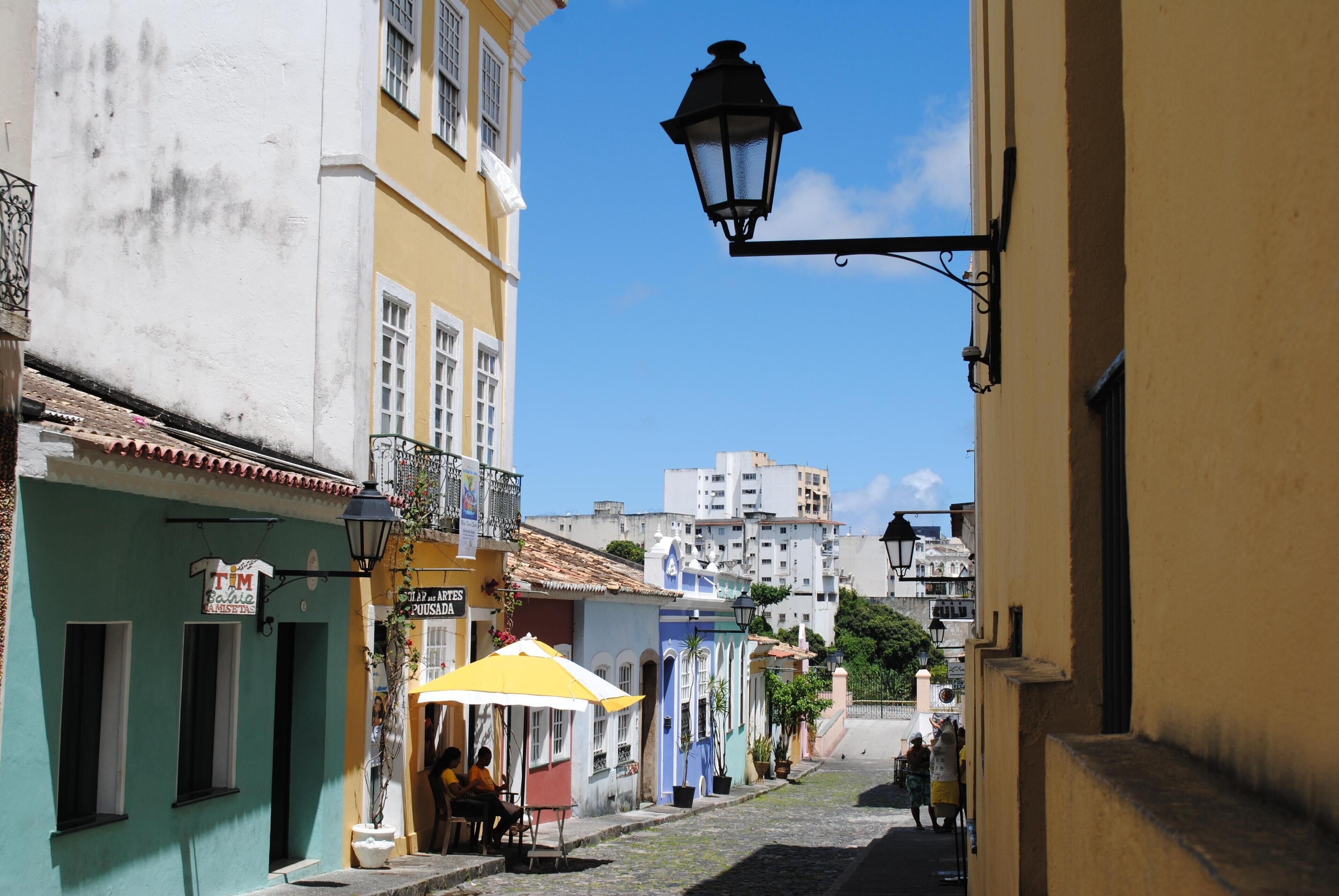 Une ruelle étroite pavée de pierres, bordée de bâtiments aux couleurs vives, un café avec des tables en terrasse et un lampadaire.