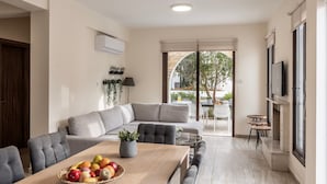 Ein modernes Wohnzimmer mit einem grauen Ecksofa, einem Holzesstisch mit Obst und einem an der Wand montierten Fernseher.