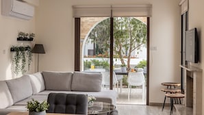 Ein modernes Wohnzimmer mit einem grauen Sofa, einem Couchtisch und einem an der Wand montierten Fernseher. Eine Glasschiebetür führt zu einem Außenbereich mit Tisch und Stühlen.
