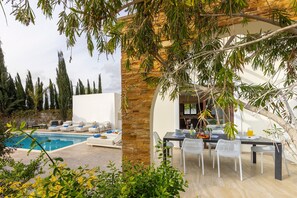 Un área junto a la piscina con una mesa y sillas, rodeada de vegetación y un muro de piedra.