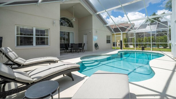 A covered outdoor swimming pool with lounge chairs and a table, surrounded by a house with large windows and a patio area. - Amazing 6 Bedrooms With Pool (Kissimmee)