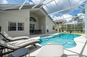 A covered outdoor swimming pool with lounge chairs and a table, surrounded by a house with large windows and a patio area. - Amazing 6 Bedrooms With Pool (Kissimmee)