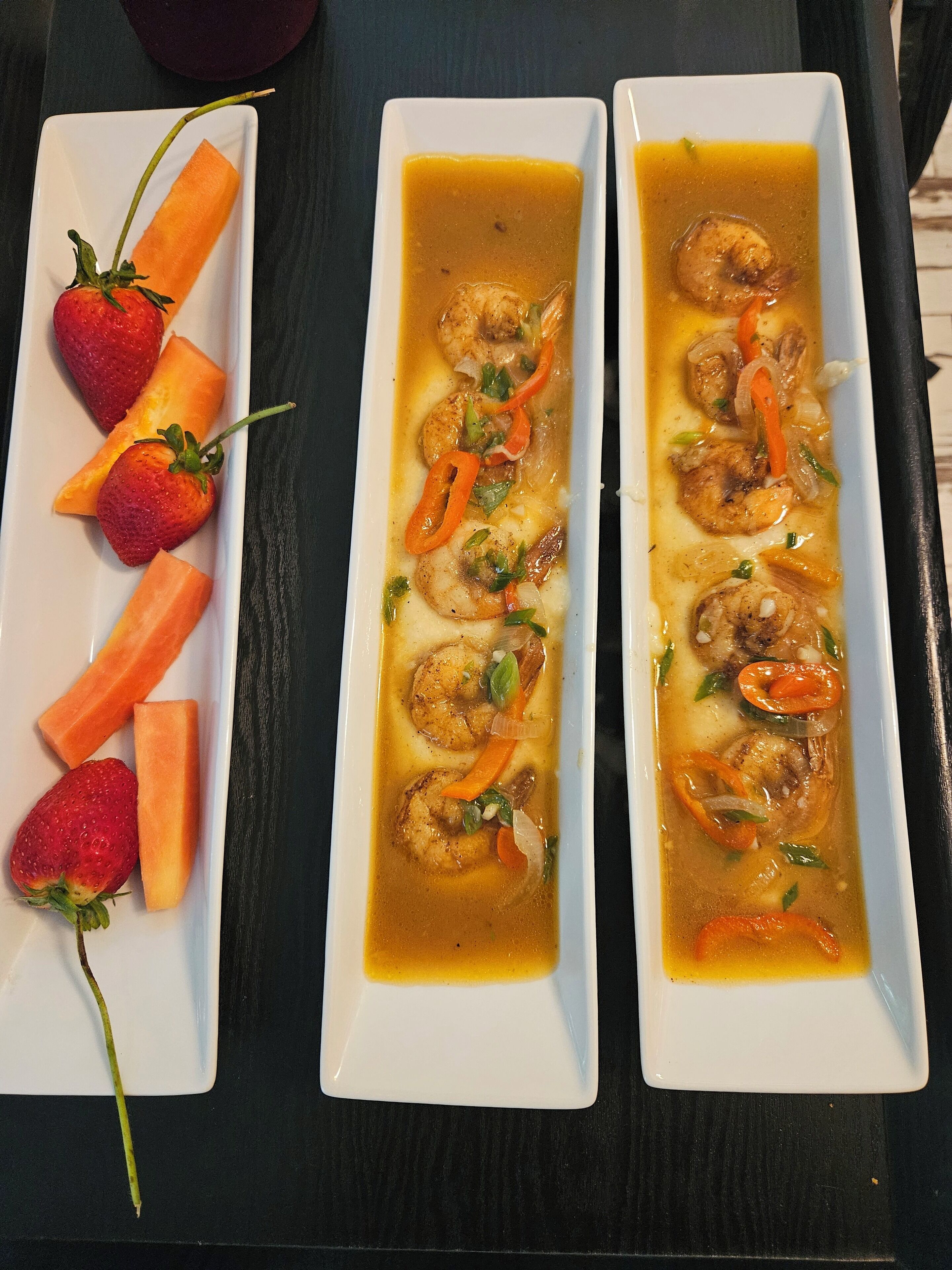 Two rectangular white dishes with shrimp and vegetables in a broth, accompanied by sliced strawberries and carrots on a dark surface.