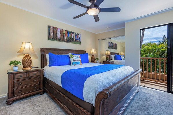 Une chambre à coucher comprenant un lit, des tables de chevet, un ventilateur de plafond, un grand miroir et un balcon avec vue sur la verdure.