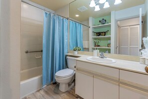 Une salle de bain avec une baignoire, des toilettes et un lavabo. On y trouve un rideau de douche bleu, un miroir au-dessus du lavabo et une étagère avec des objets décoratifs.