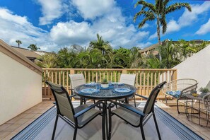 Une table ronde entourée de chaises, avec une petite plante en pot, sur une terrasse offrant une vue sur des palmiers et une clôture en bois.
