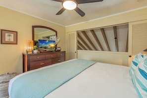 Une chambre à coucher comprenant un lit, une commode, un ventilateur de plafond, un grand miroir et une fenêtre avec des stores.