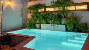 Ein Schwimmbecken mit Stufen, umgeben von Topfpflanzen und einem Gebäude mit großen Fenstern sowie einem Turm im Hintergrund.