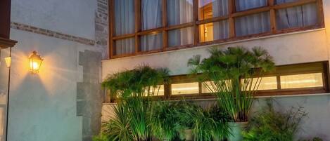 Una piscina con escalones de acceso, rodeada de macetas con plantas y un edificio con grandes ventanales y una torre al fondo.