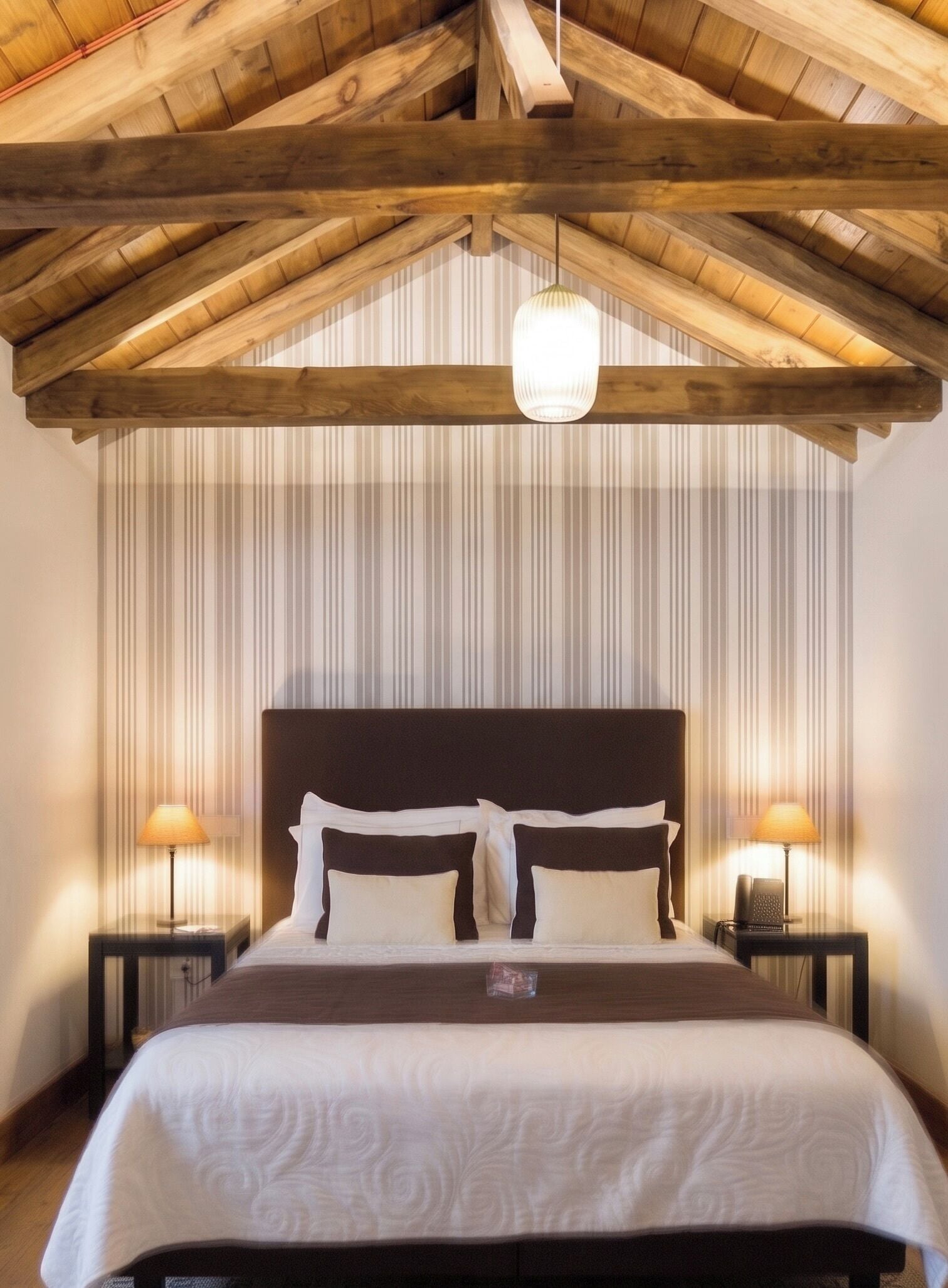 Ein Schlafzimmer mit Holzbalkendecke, einem Bett mit weißer Bettwäsche, zwei Nachttischlampen und einer gestreiften Wand.