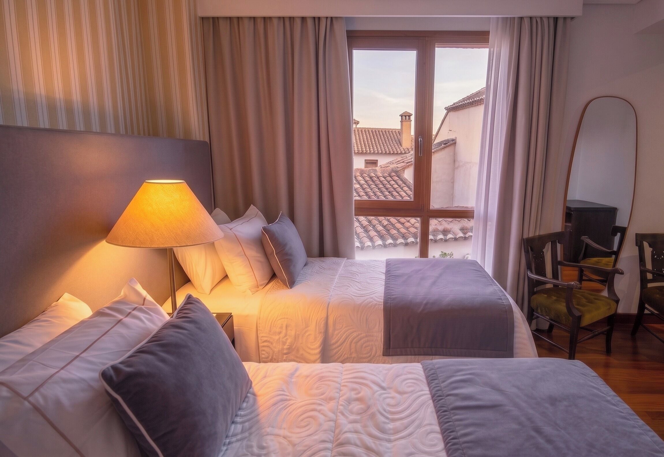A hotel room with a bed, a lamp, a chair, and a view of buildings through a window.