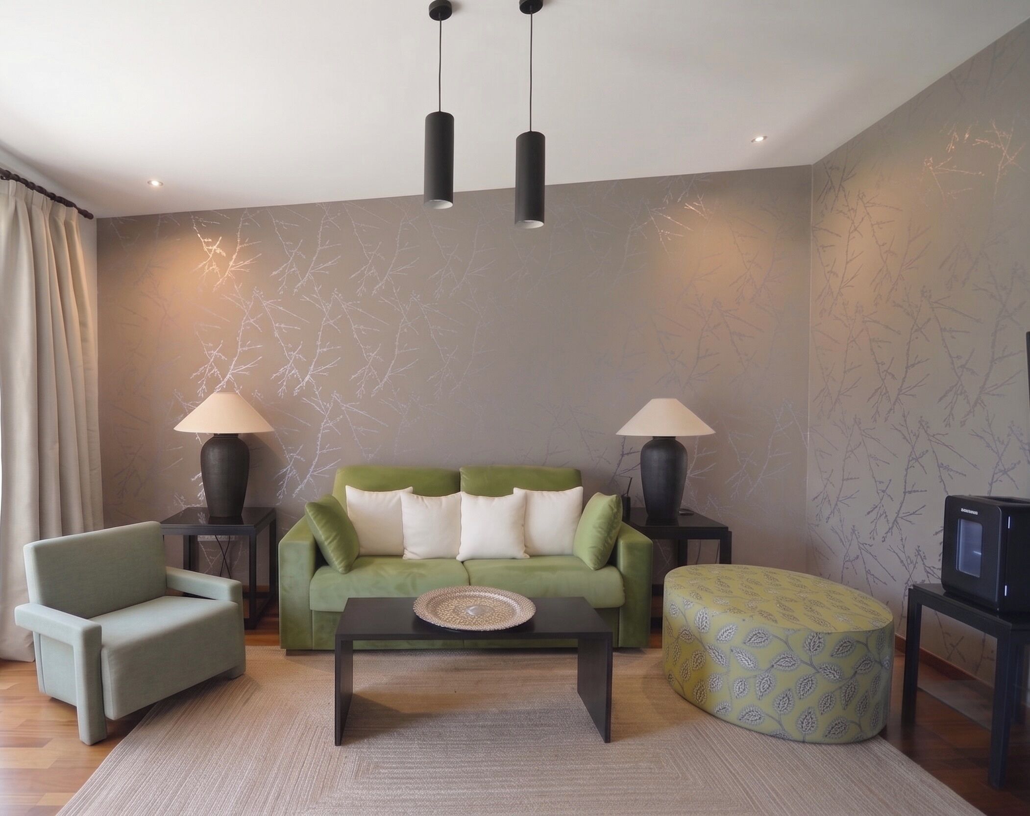 Een woonkamer met een groene bank, twee bijzettafels met lampen, een salontafel, een ottoman met patroon en een televisietoestel op een meubel.