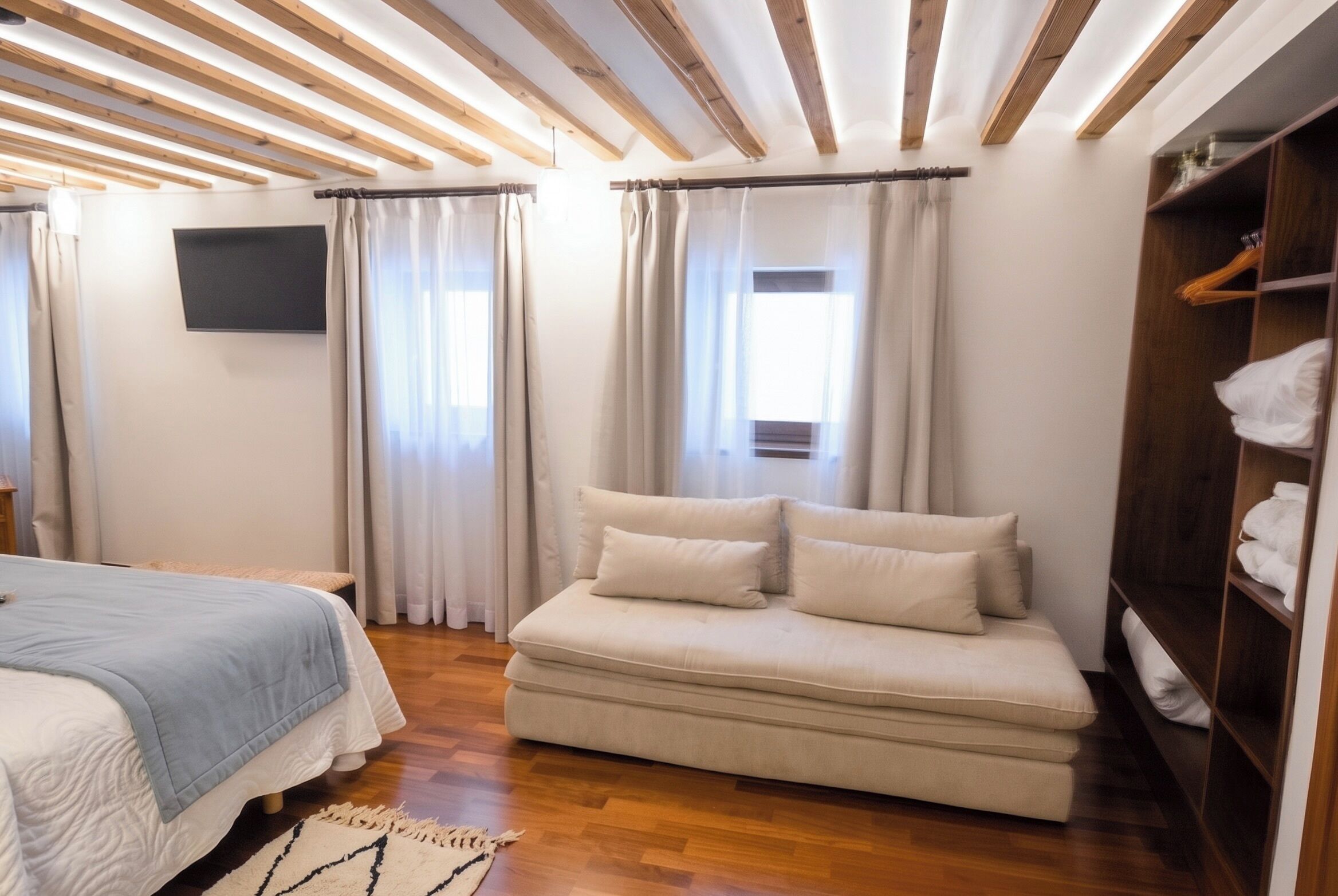 A bedroom with a bed, a sofa, a TV, wooden beams, a wardrobe, and a window.