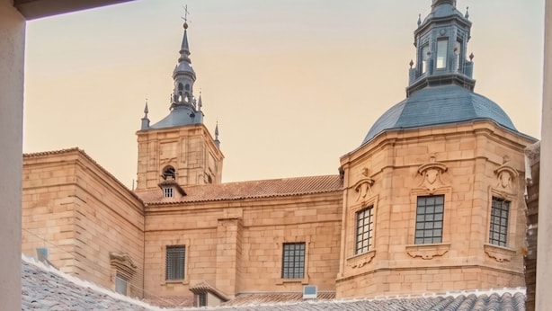 Ein Innenhof mit einem Gebäude, das eine Kuppel und einen Glockenturm sowie einen hölzernen Balkon und leuchtend rote Blumen im Vordergrund zeigt.