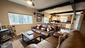 Un salon avec un canapé en cuir marron, une table basse et une cheminée. Au fond, on aperçoit une cuisine avec des meubles en bois et un coin repas.