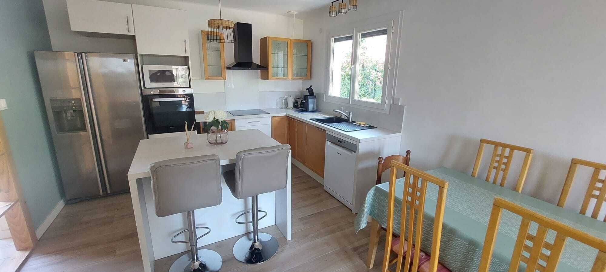 A modern kitchen with a central island, bar stools, wooden chairs, a dishwasher, and a window providing natural light.