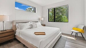 Ein modernes Schlafzimmer mit einem großen Bett, zwei Nachttischen, einem Beistelltisch, einem Sessel und zwei Fenstern mit Blick ins Freie.