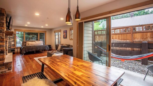 Un grand espace de vie comprenant une grande table en bois, un canapé et un lit. À l’extérieur, on trouve un hamac et, sur la gauche, un mur en pierre.