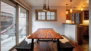 Un coin repas avec une table et des chaises en bois, une grande fenêtre avec des stores et un espace cuisine doté de meubles en bois et d’un éclairage suspendu.