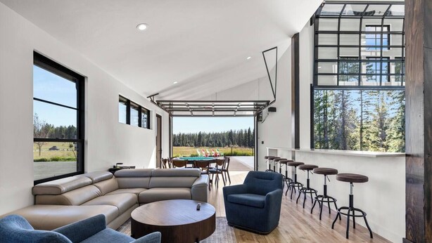 A spacious living area with a large L-shaped sofa, a round coffee table, and a blue armchair. There are multiple bar stools along the wall and a view of a deck area with outdoor seating. - Paddles Up Private Pickleball Retreat (Whitefish)