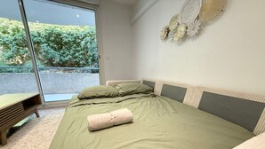 A bedroom with a bed, pillows, a bench, and a view of greenery through a sliding door.