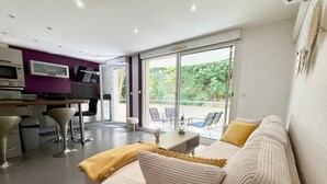 A modern living room with a sofa, a coffee table, and a dining area with chairs. There is a kitchen area with appliances and a TV. The room has large windows with a view of greenery outside.