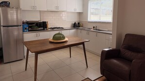Una cocina moderna con armarios blancos, un refrigerador de acero inoxidable, un microondas y una estufa a gas. Hay una mesa de madera con una cubierta verde abovedada y una silla de cuero marrón.