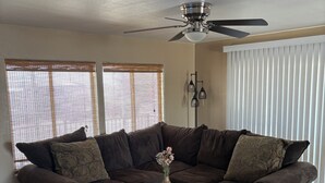 Een woonkamer met een hoekbank, een kleine ronde tafel met een vaas, en een plafondventilator.