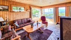 L'interno accogliente di una baita con un divano in tessuto tartan, un tavolino in legno e una poltrona rossa. Ampie finestre offrono vista sul panorama circostante.