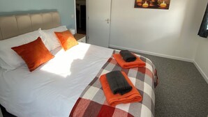A neatly made bed with a checkered blanket, two orange pillows, and a folded towel. There is a painting of candles on the wall, a ceiling light fixture, and a door leading to another room.