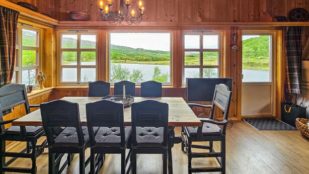 A dining area with a wooden interior, large windows offering a view of a lake and hills, and a chandelier. - Cozy home in Hemsedal with sauna (Hemsedal)