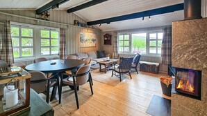 Un salon chaleureux avec une cheminée, un plancher en bois et de grandes fenêtres donnant sur l’extérieur.